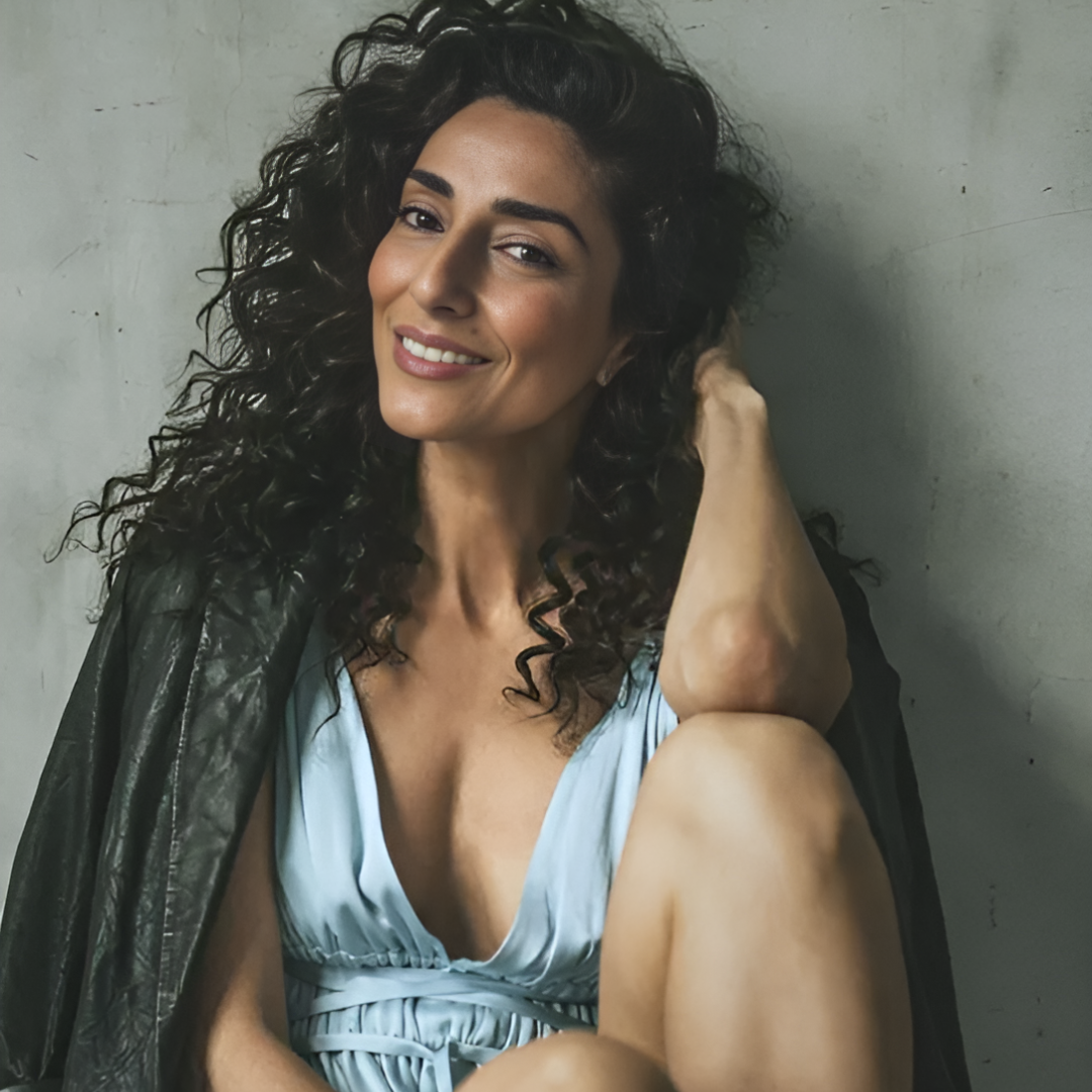 Necar Zadegan shown sitting against a grey wall in a blue dress and black leather jacket